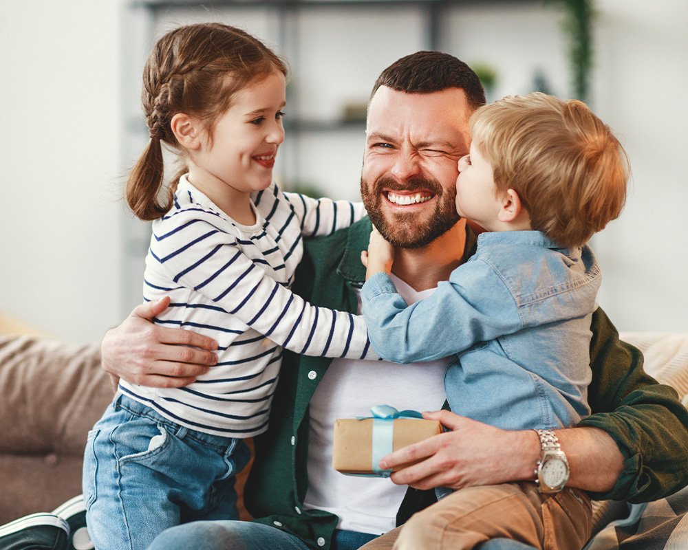 Vater umarmt zwei Kinder und hält ein Geschenk in seinen Händen