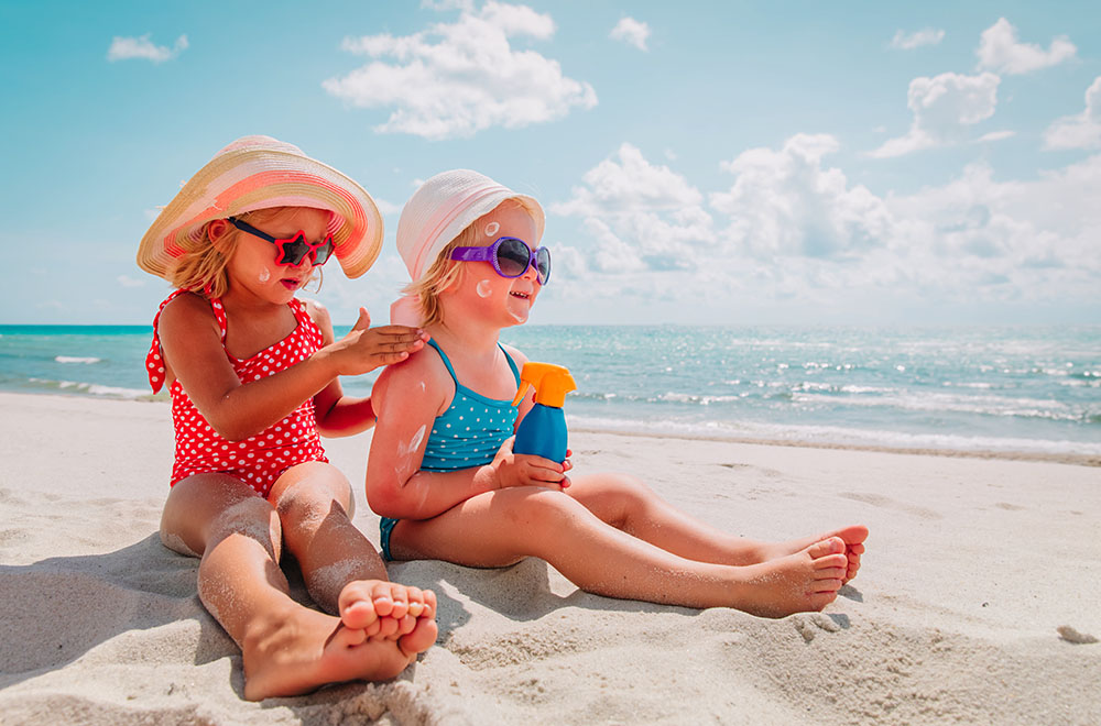 Zwei Mädchen in Badeanzügen, mit Schwimmbrillen und Hüten am Strand, beim Sonnencremen.