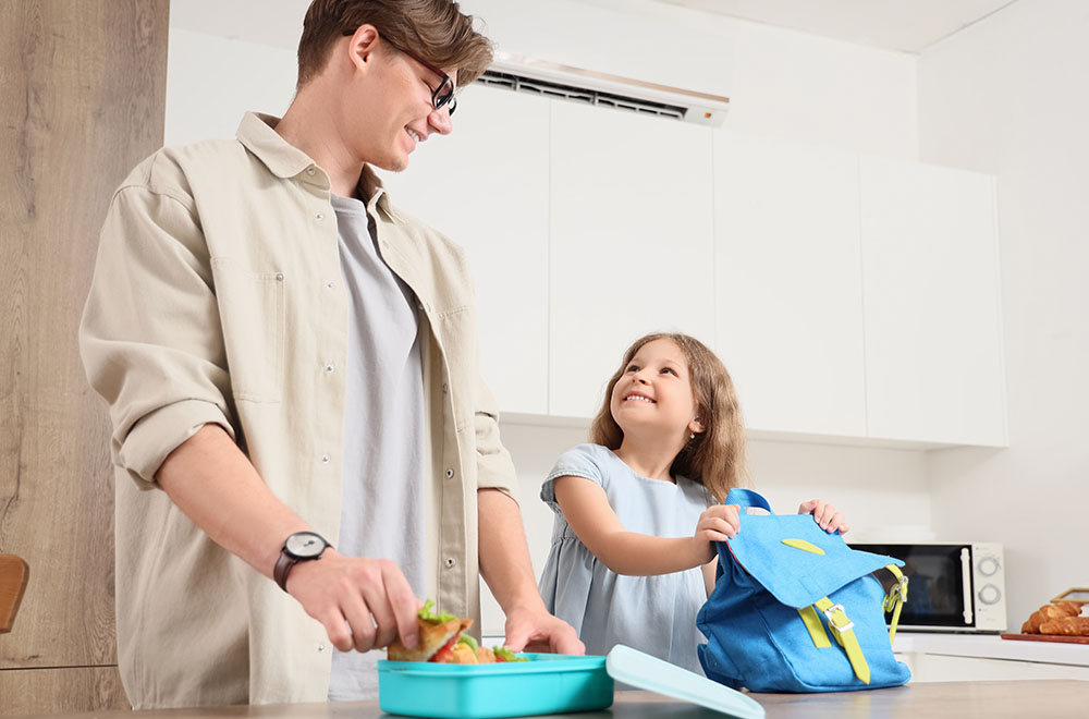 Der Vater bereitet das Mittagessen für seine Tochter vor, die lächelnd ihren Rucksack hält.