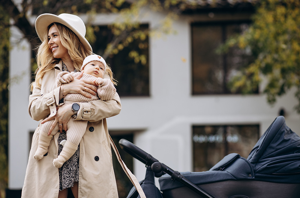 Eine Frau hält ein Baby im Arm und geht neben einem Kinderwagen vor einem Gebäude.